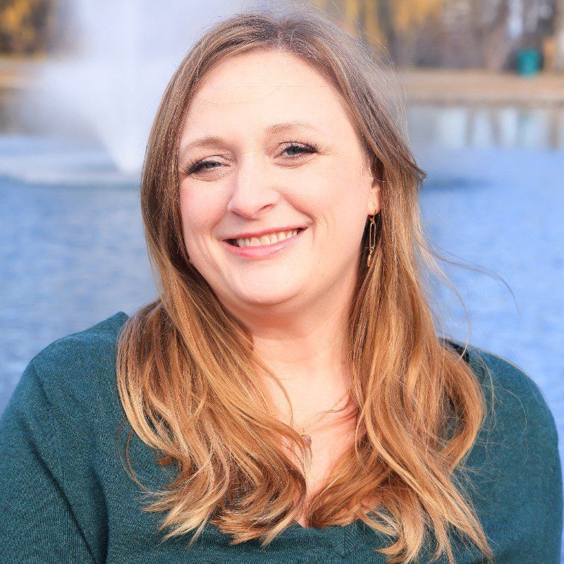 A woman with long, light brown hair smiles at the camera. She is wearing a dark green sweater, and the background shows a blurred fountain in a body of water.