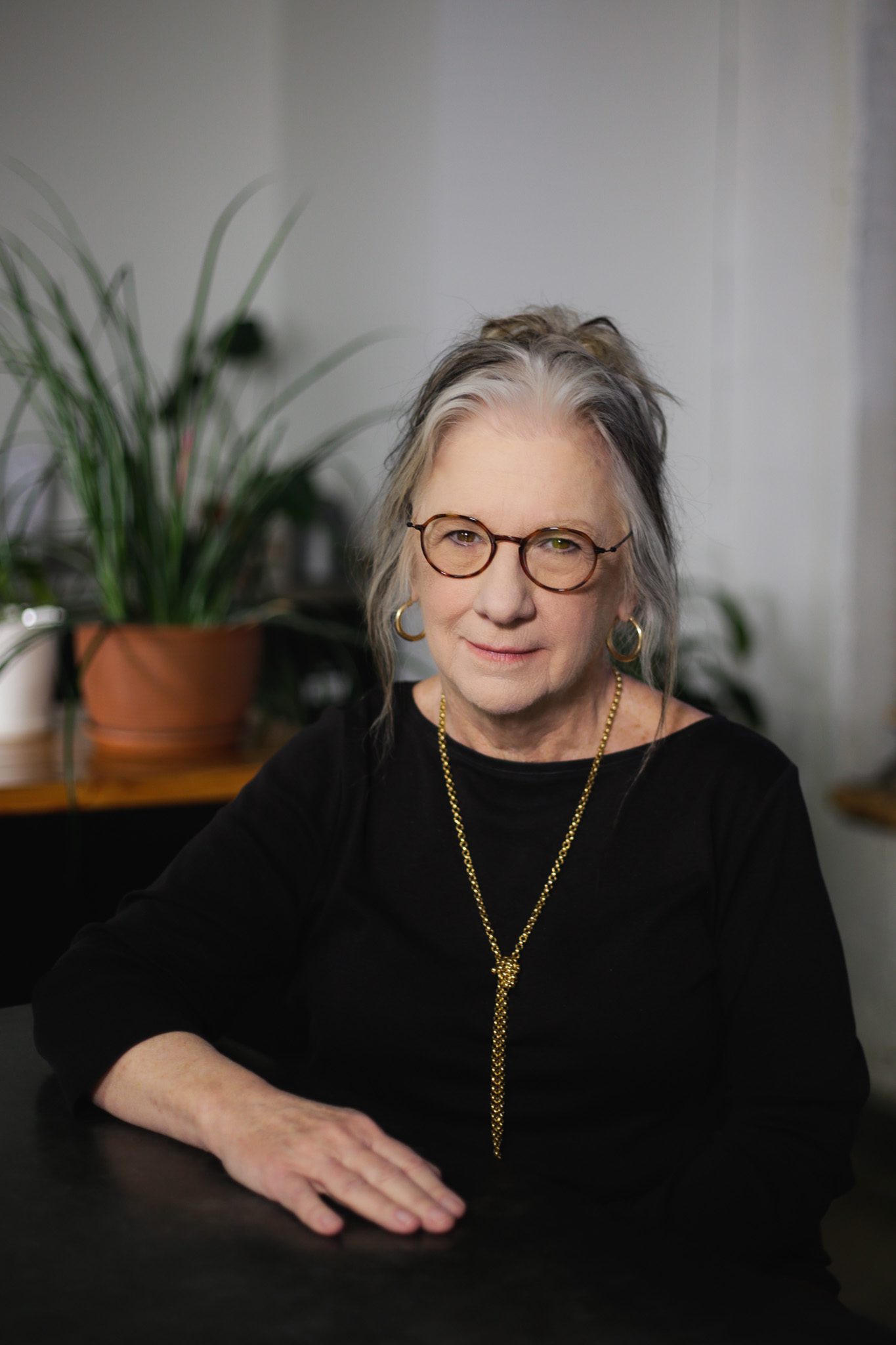 Profile picture of an older woman with glasses, long gray hair tied back, wearing a black top, gold necklace, and earrings, seated in front of a potted plant