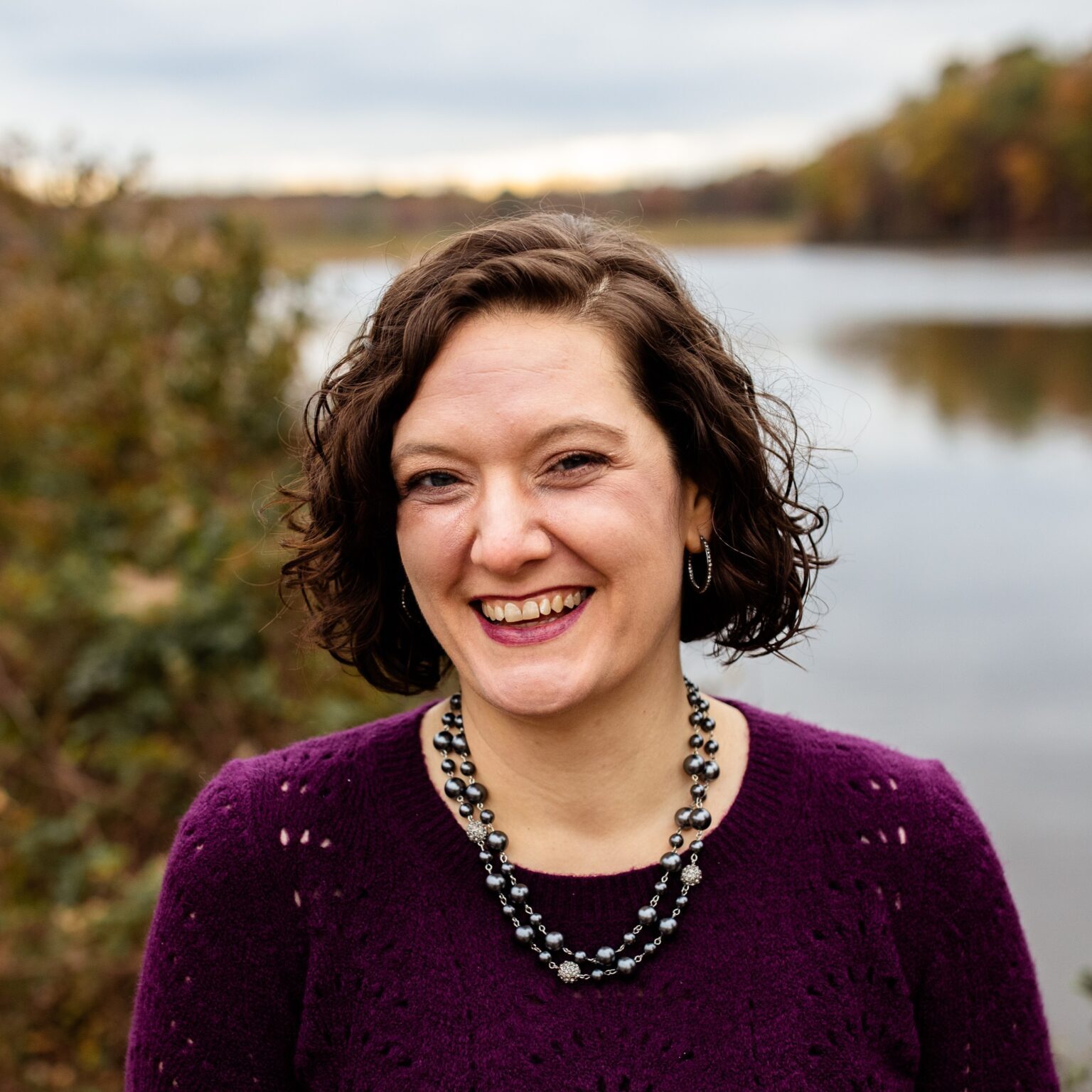 A woman with short, curly brown hair smiles broadly at the camera. She is wearing a purple sweater and a layered bead necklace. The background shows a calm river and autumn trees.