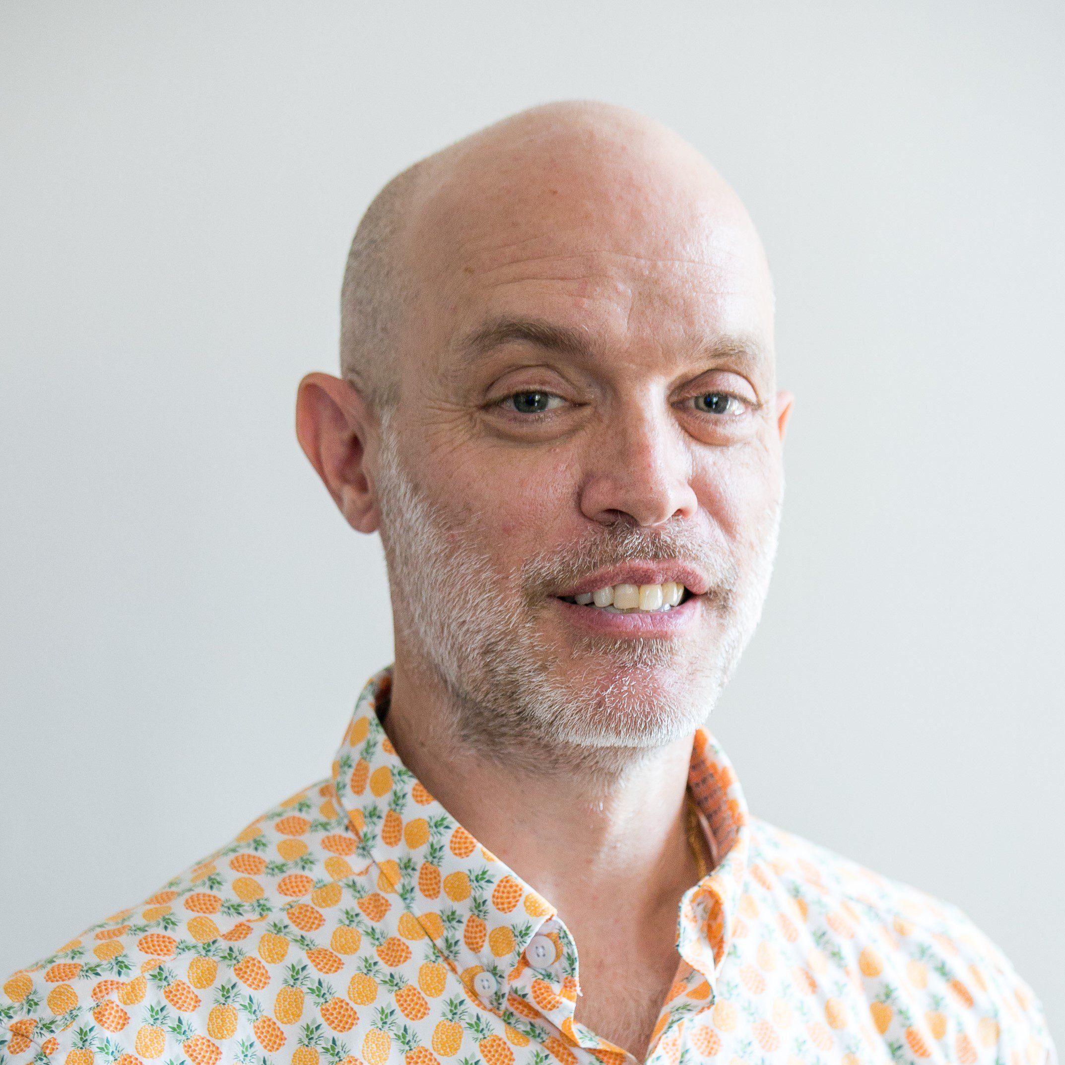 A man with a bald head and a short beard smiles slightly. He is wearing a light orange shirt with a pineapple print.