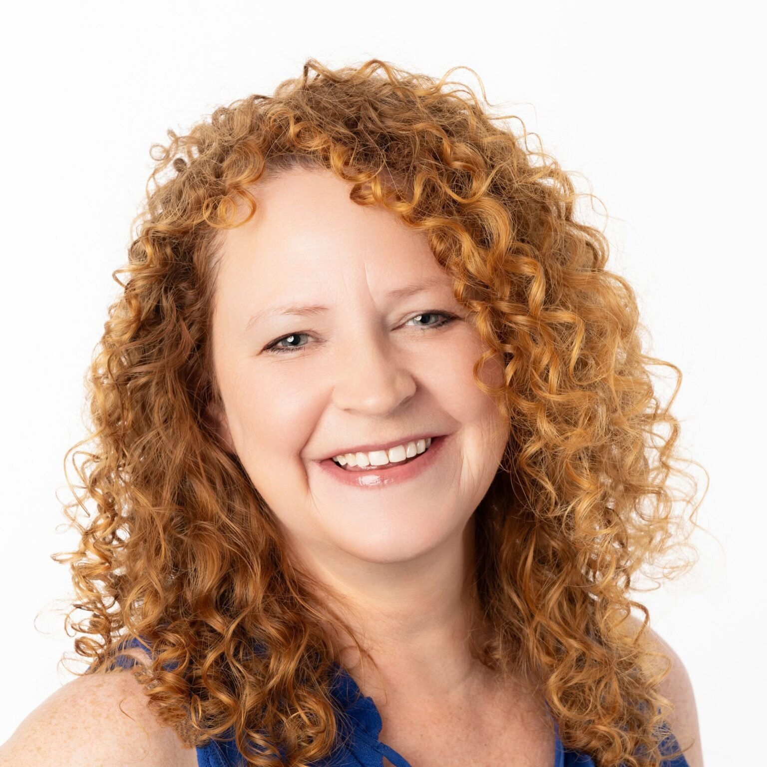 A woman with curly red hair smiles at the camera. She is wearing a sleeveless blue top.