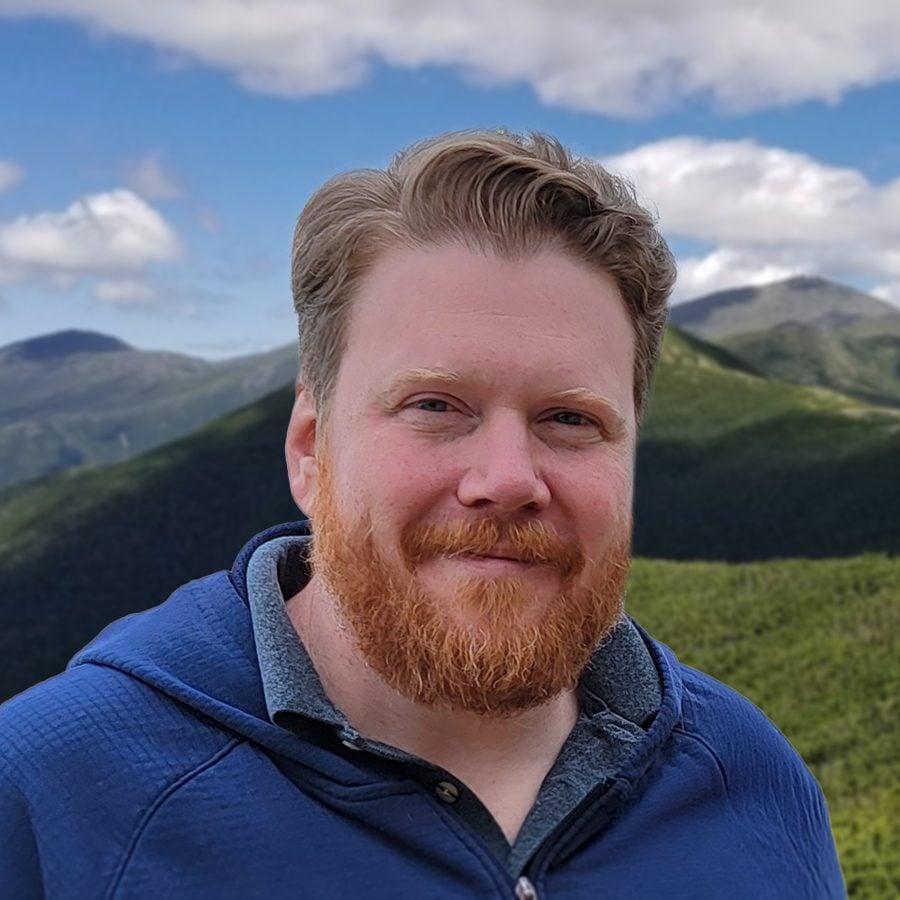 A man with short, light brown hair and a red beard smiles slightly at the camera. He is wearing a blue jacket, and the background shows green mountains under a partly cloudy sky.