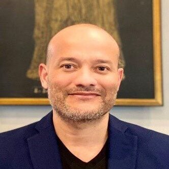 A bald man with a light beard smiles at the camera. He is wearing a dark blue blazer over a black shirt.