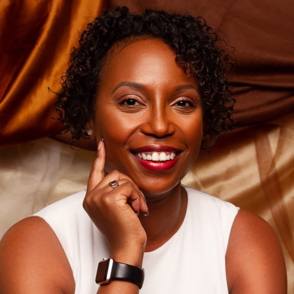 A smiling woman with short curly hair wearing a white top and a black smartwatch. She rests her chin on her hand, with a ring visible on her finger. The background features brown and beige draped fabric.