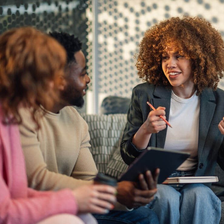 Three people engaged in a discussion while sitting on a couch. The central figure, a woman with curly hair, is animatedly talking and holding a pen and a clipboard. The others, a man and a woman, are listening attentively.