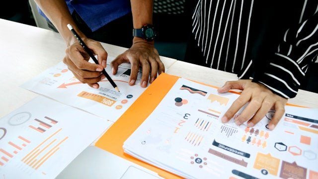 Two people reviewing documents with charts and graphs on a desk, one person pointing with a pen. The documents contain various types of data visualizations in orange and black colors.